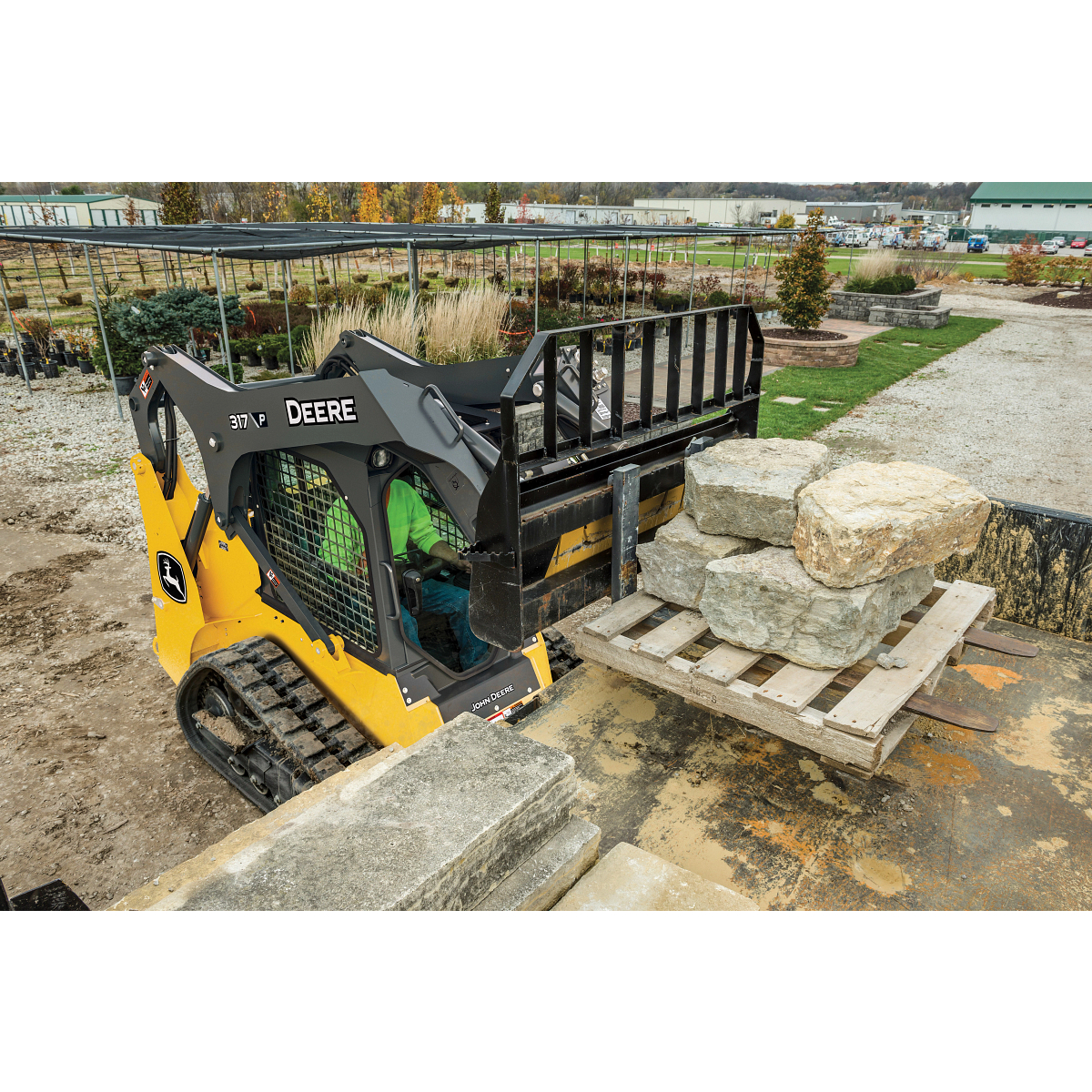 A compact track loader using a pallet fork to lift a wooden pallet stacked with large stone blocks in a landscaping yard