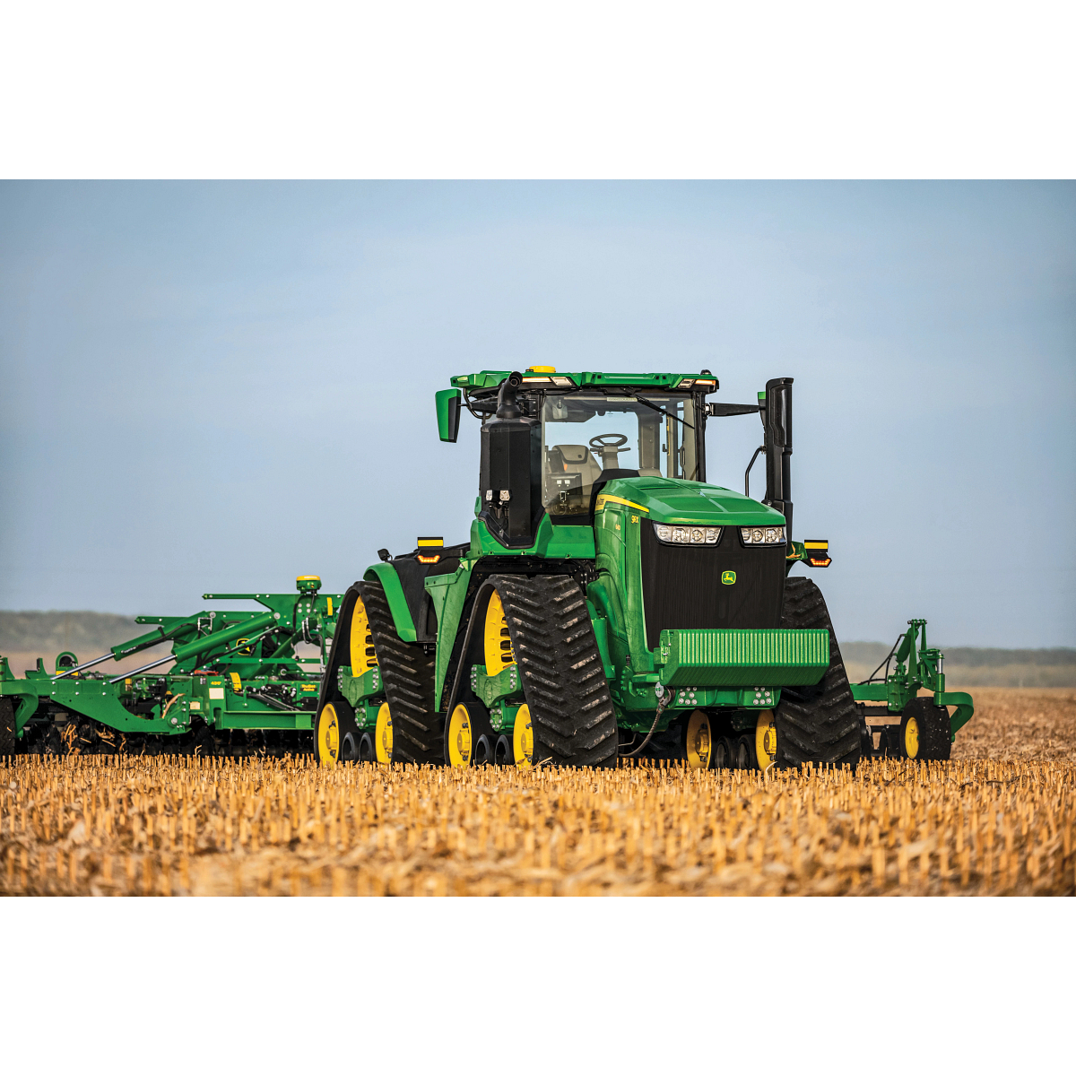 Front view of a large green tracked tractor with a tillage implement hitched to the rear sitting in a harvested field covered with corn stubble under a clear blue sky