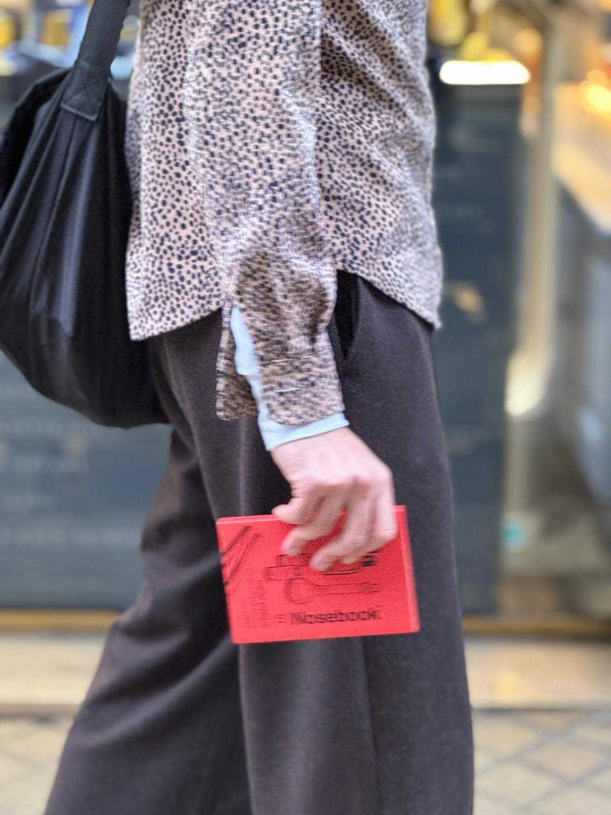 Person portant un haut imprimé léopard et un pantalon marron foncé tenant un coffret rouge Nosebook en marchant.