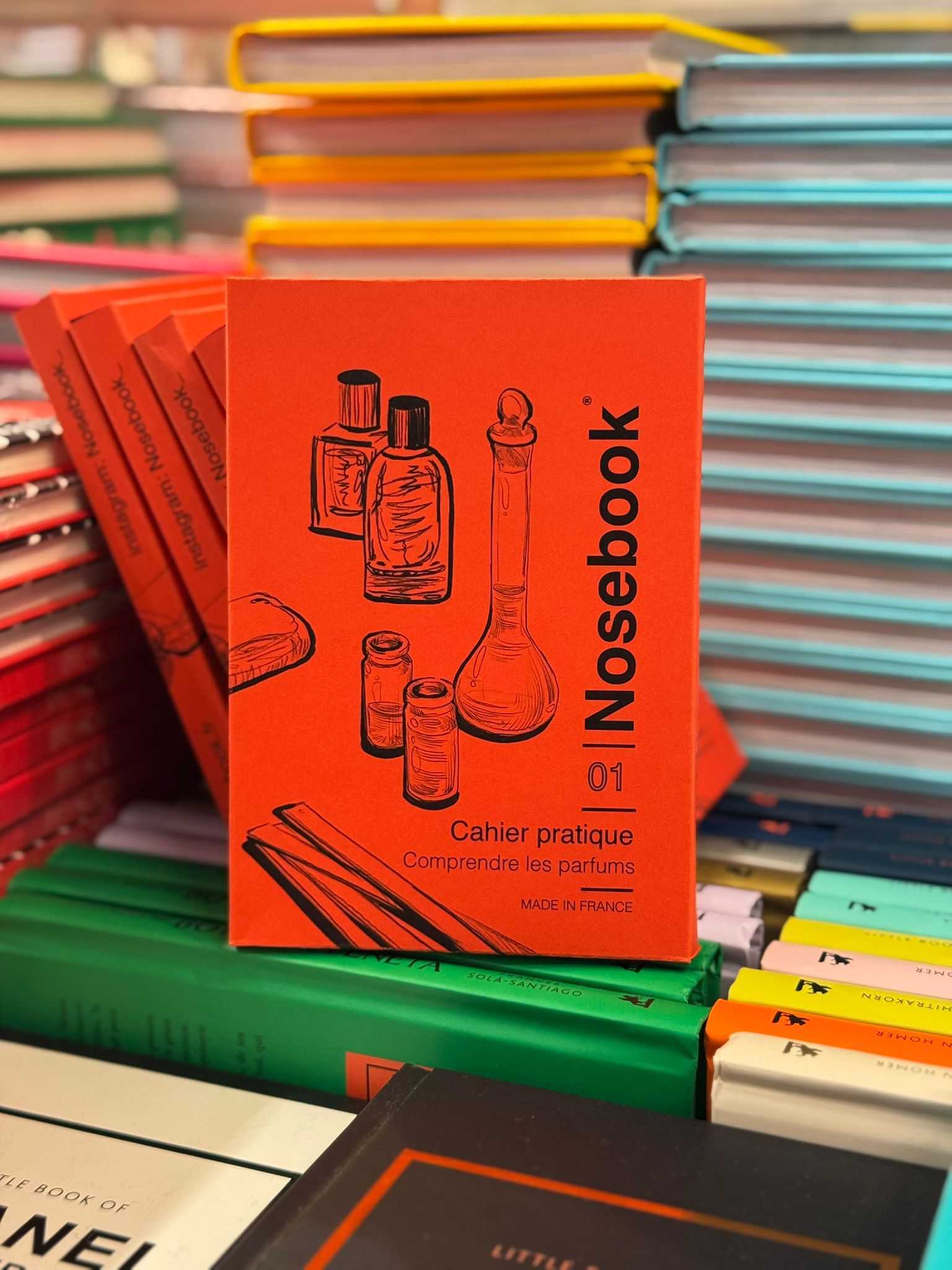 Plusieurs exemplaires du carnet rouge-oranger, 'Nosebook 01 Cahier pratique Comprendre les parfums', posés devant une pile de livres colorés.