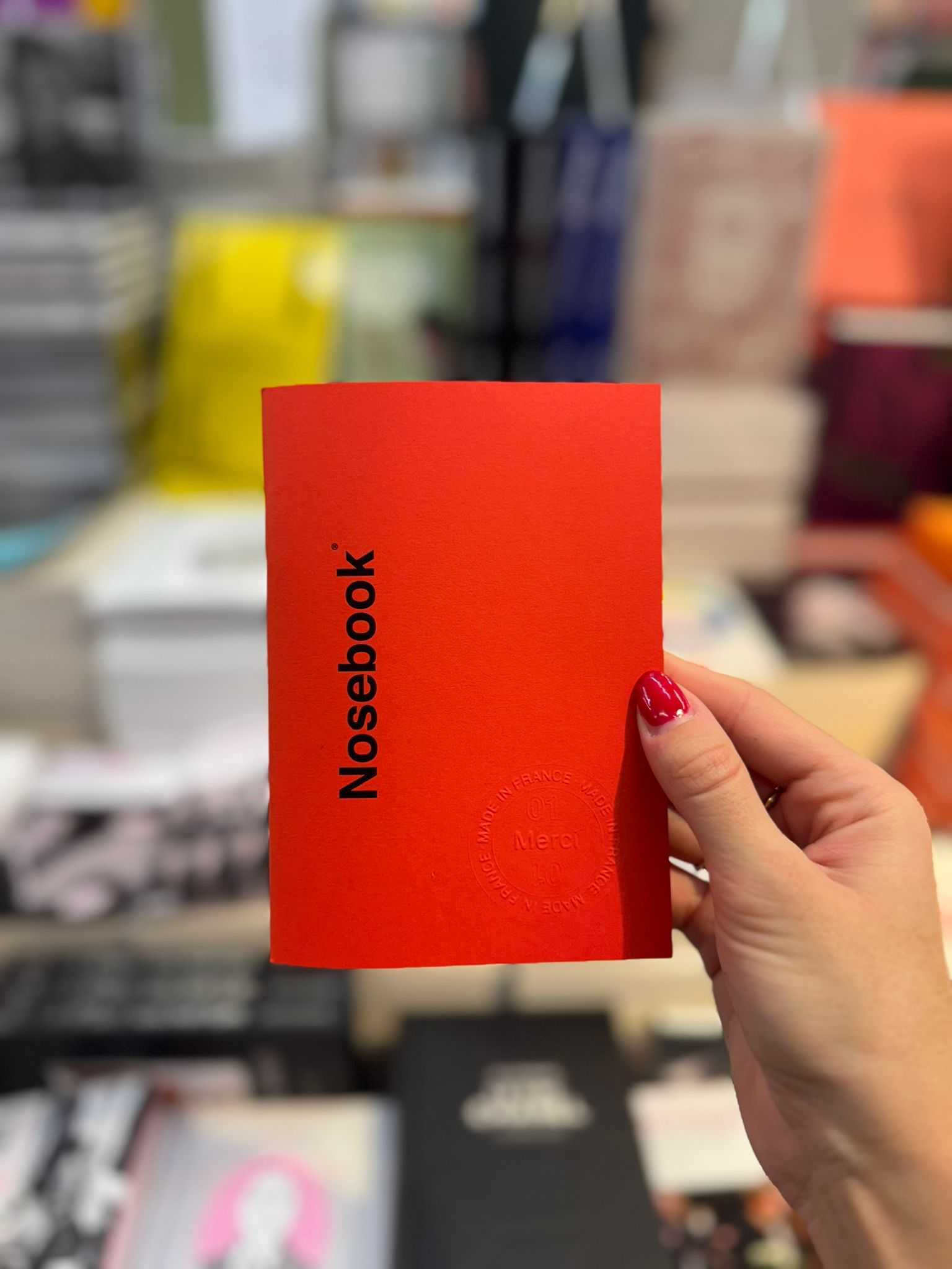 Main droite tenant un carnet rouge avec l'inscription noire "Nosebook" et un sceau embossé, made in France au tour et merci au centre.
