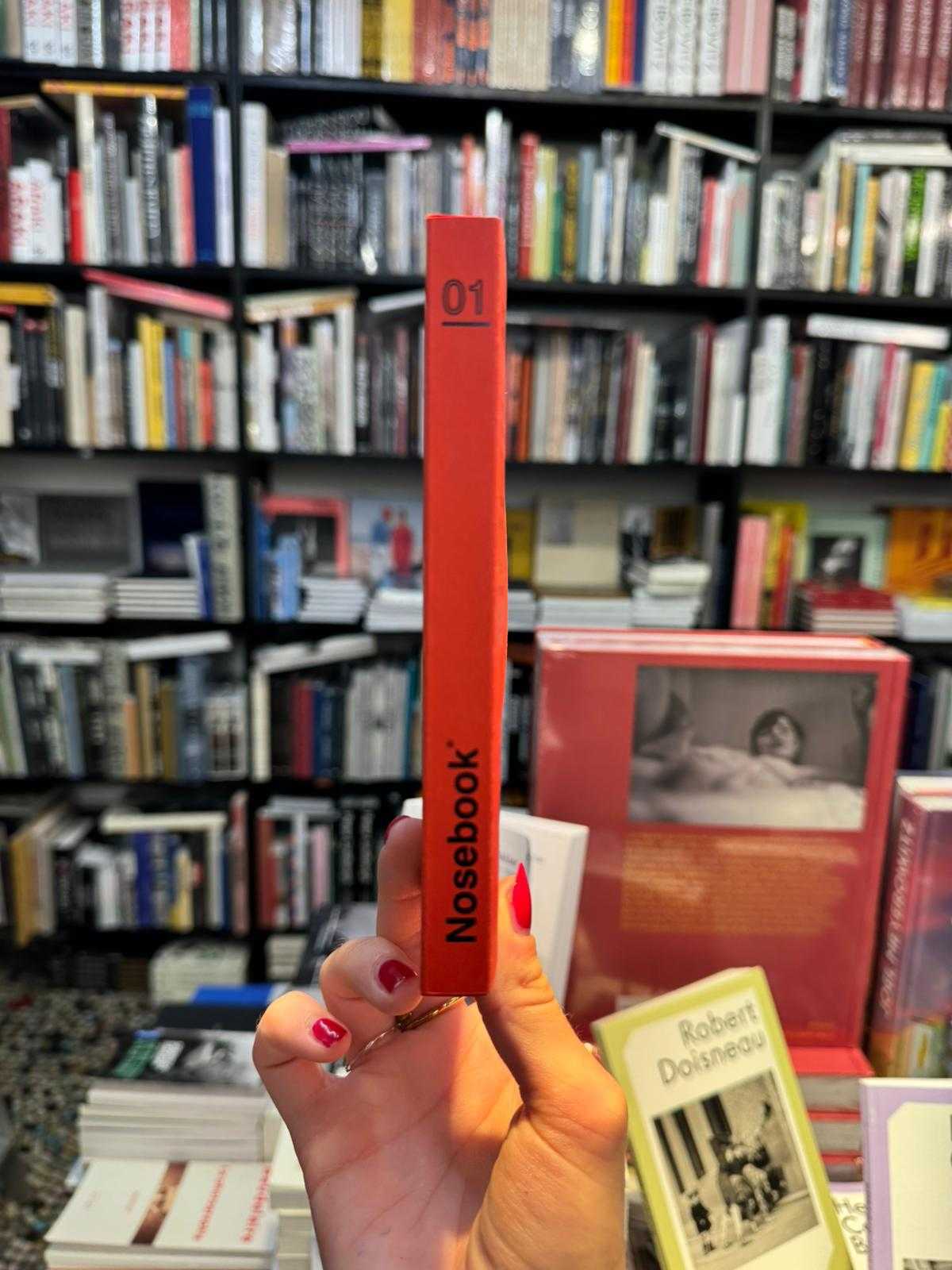 Main de femme tenant un livre rouge avec le titre 'Nosebook' et le numéro 01 sur la tranche, devant des étagères remplies de livres.