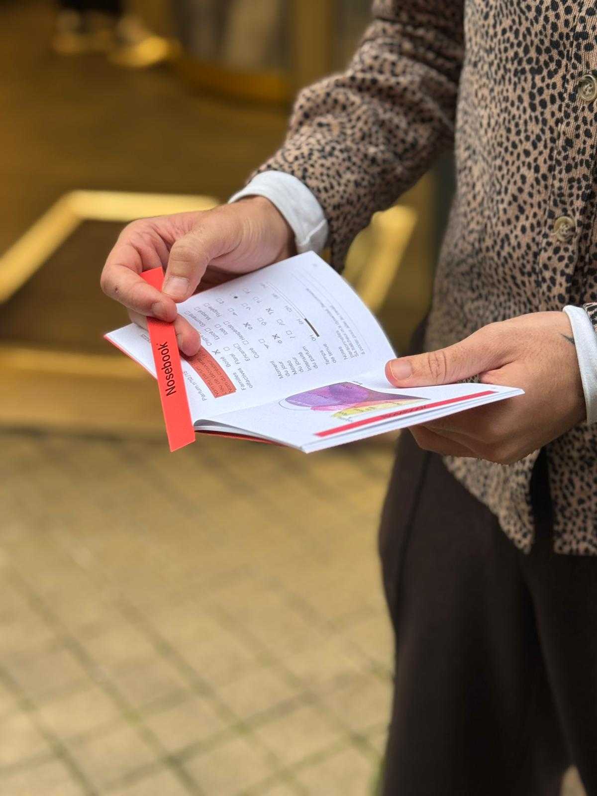 Person tenant un petit livre ouvert sur une page avec un diagramme expliquant l'évolution d'un parfum et son analyse (telle que la famille olfactive, la météo du jour, l'intensité et la teneur de ce dernier), avec une touche rouge portant l'inscription 'Nosebook'.
