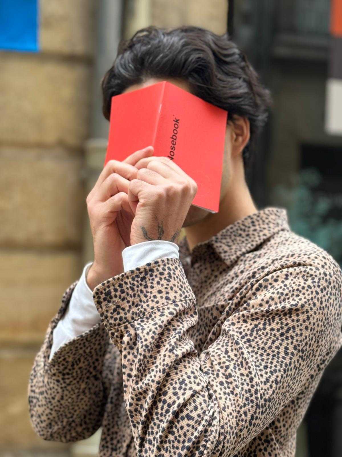 Person portant une chemise à motifs léopard tient un petit livre rouge, nommé Nosebook, couvrant son visage.