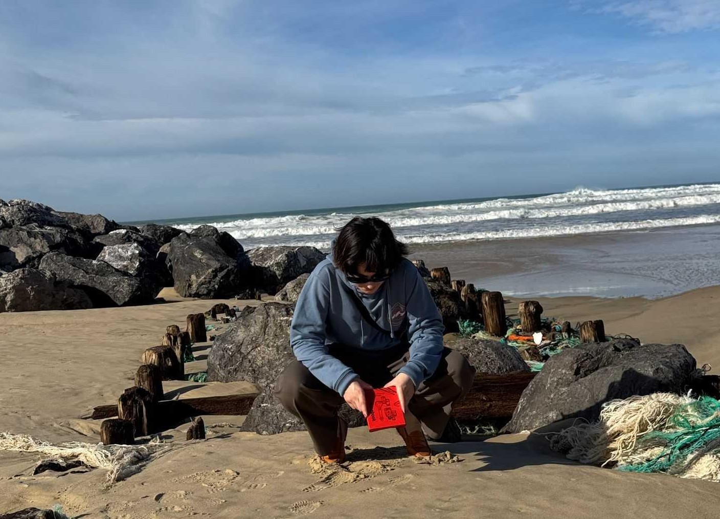 Une personne accroupie sur une plage sablonneuse tenant un livret rouge, Nosebook avec l'océan et des rochers en arrière-plan.