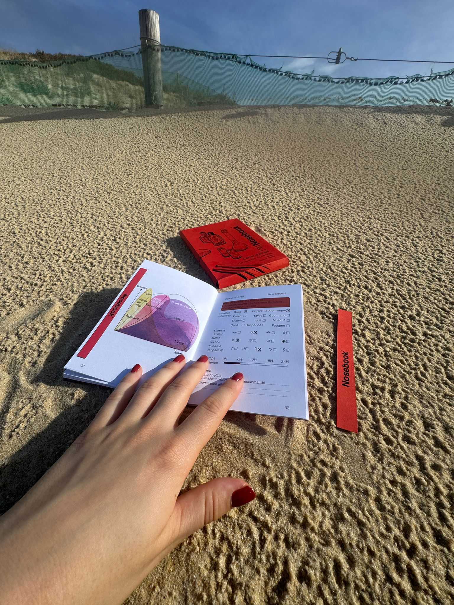 Main avec vernis rouge touchant un carnet ouvert affichant un graphique et du texte sur l'analyse d'un parfum, posé sur du sable avec un coffret fermé et une touche rouge marquée Nosebook.