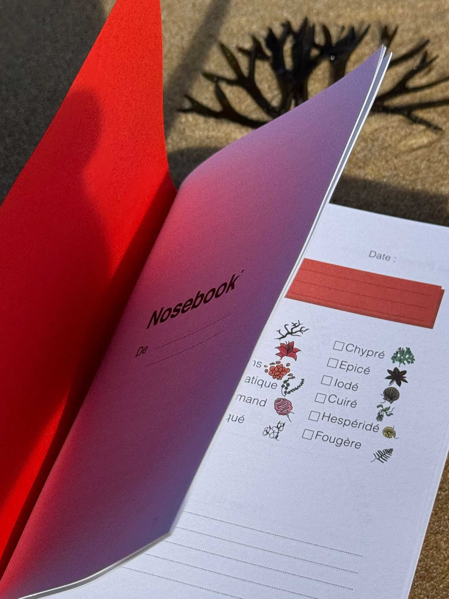 Cahier ouvert avec couverture rouge et page intérieure montrant le titre Nosebook et une liste de catégories olfactives avec cases à cocher et petites illustrations des catégories. En arière-plant une algue marine et le sable. 