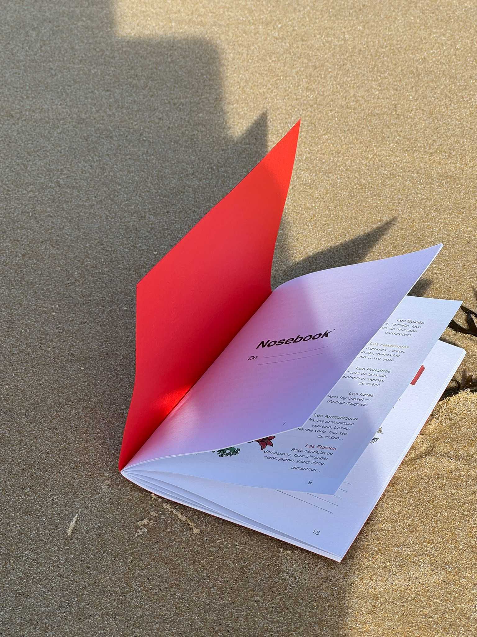 Carnet ouvert intitulé Nosebook posé sur un sol sablonneux, avec une page rouge visible et une autre page listant des épices et des arômes.