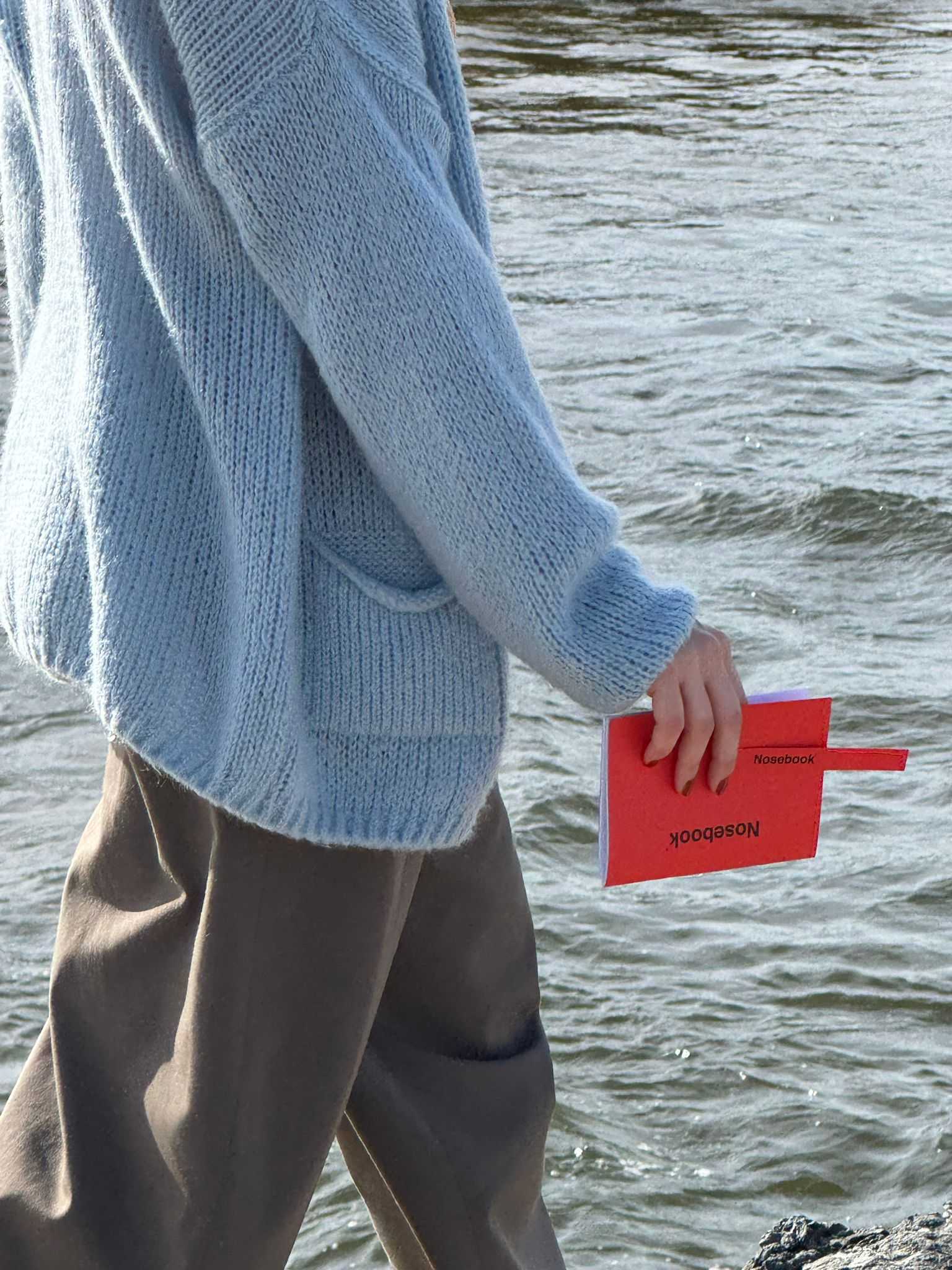 Personne portant une veste tricotée bleue et un pantalon marron ayant à la main un carnet rouge et une touche, les deux intitulés Nosebook, marchant près de l'eau.