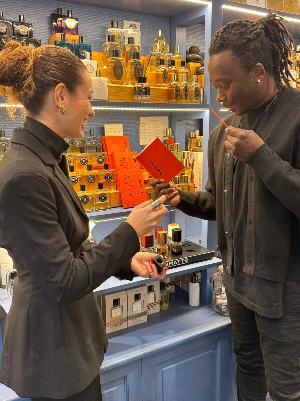 Deux personnes dans une boutique de parfums, une femme souriante tient un flacon pendant qu'un homme sent une touche de parfum et tient un Nosebook. 