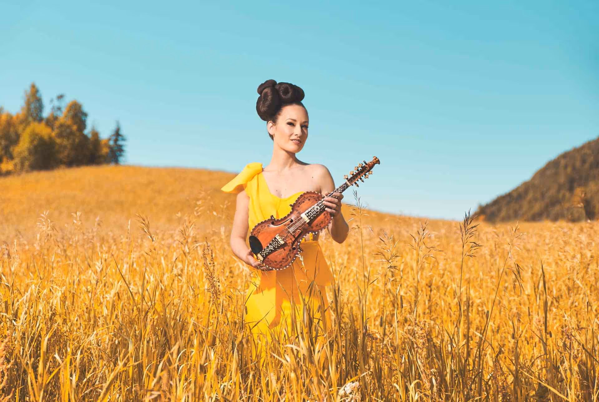 Kvinne, Ragnhild Hemsing, i gul kjole med oppsatt hår står på et åpent jorde og holder en hardingfele.