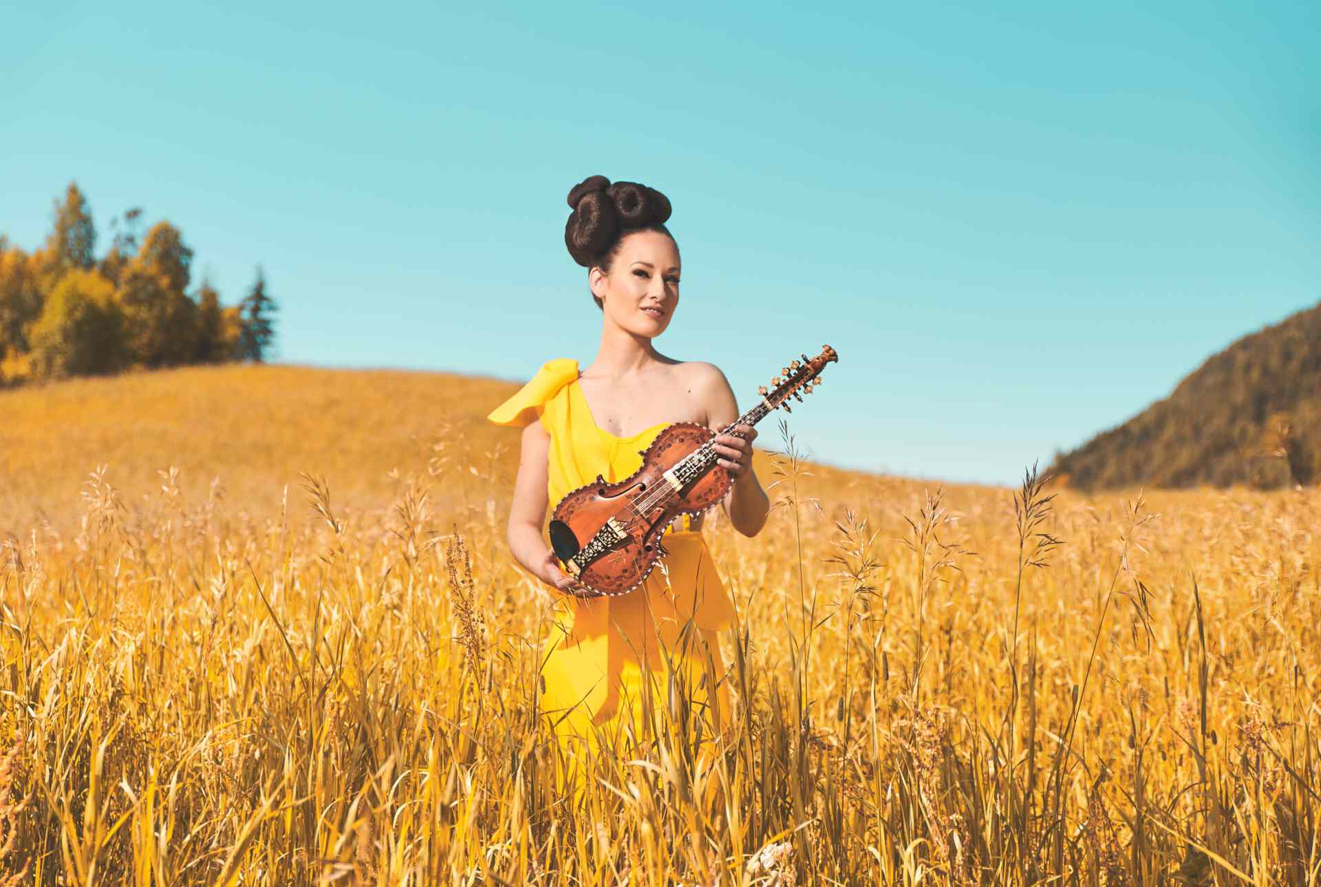 Kvinne, Ragnhild Hemsing, i gul kjole med oppsatt hår står på et åpent jorde og holder en hardingfele.