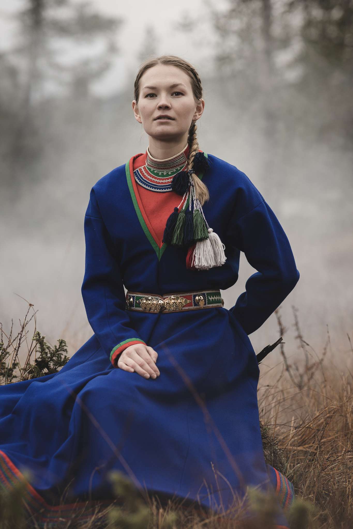 Kvinne iført tradisjonell samisk drakt med blå kåpe og dekorative dusker, sittende ute i et tåket, naturlig landskap.