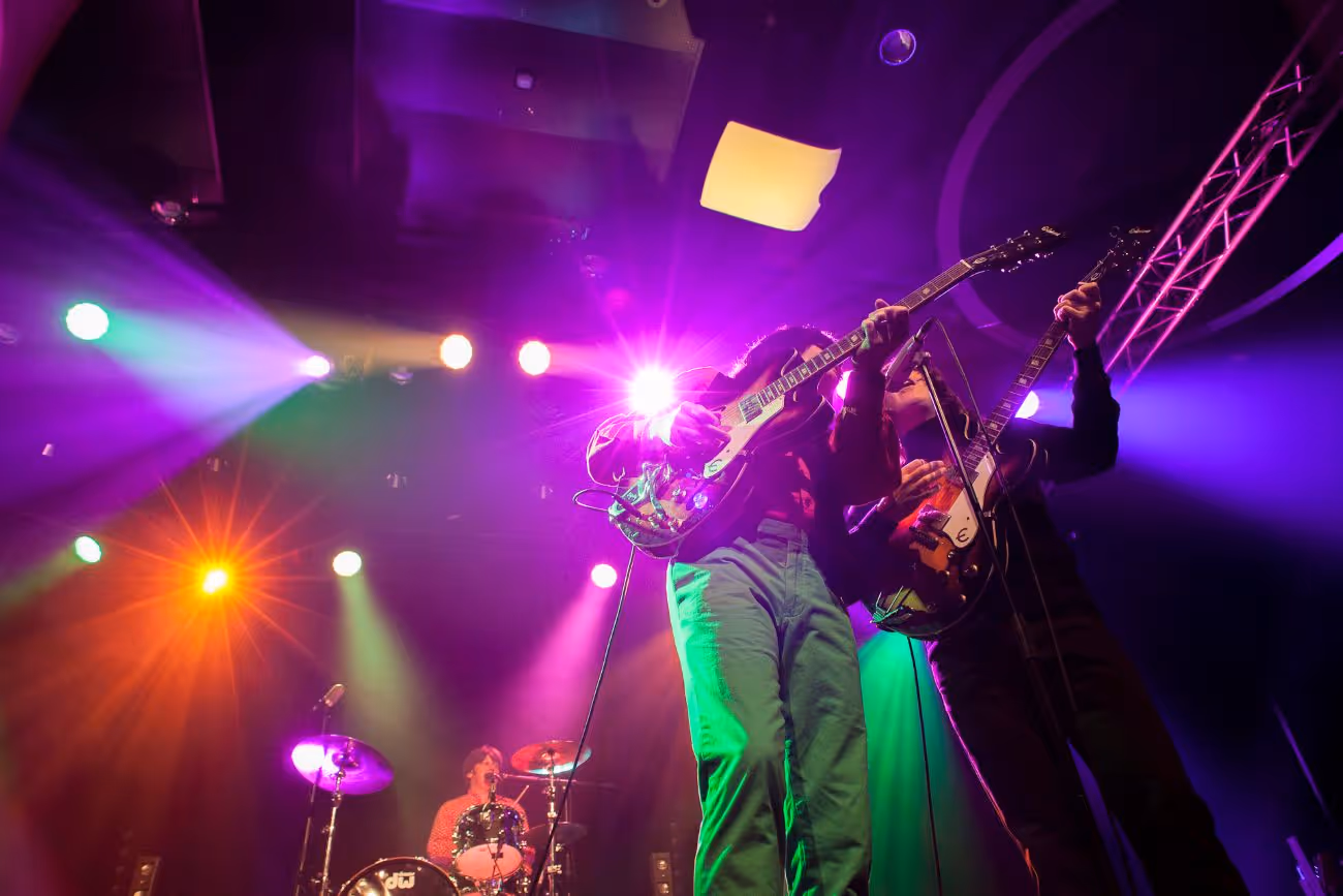 To gitarister spiller sammen på en opplyst scene med en trommeslager i bakgrunnen under fargerike konsertlys.