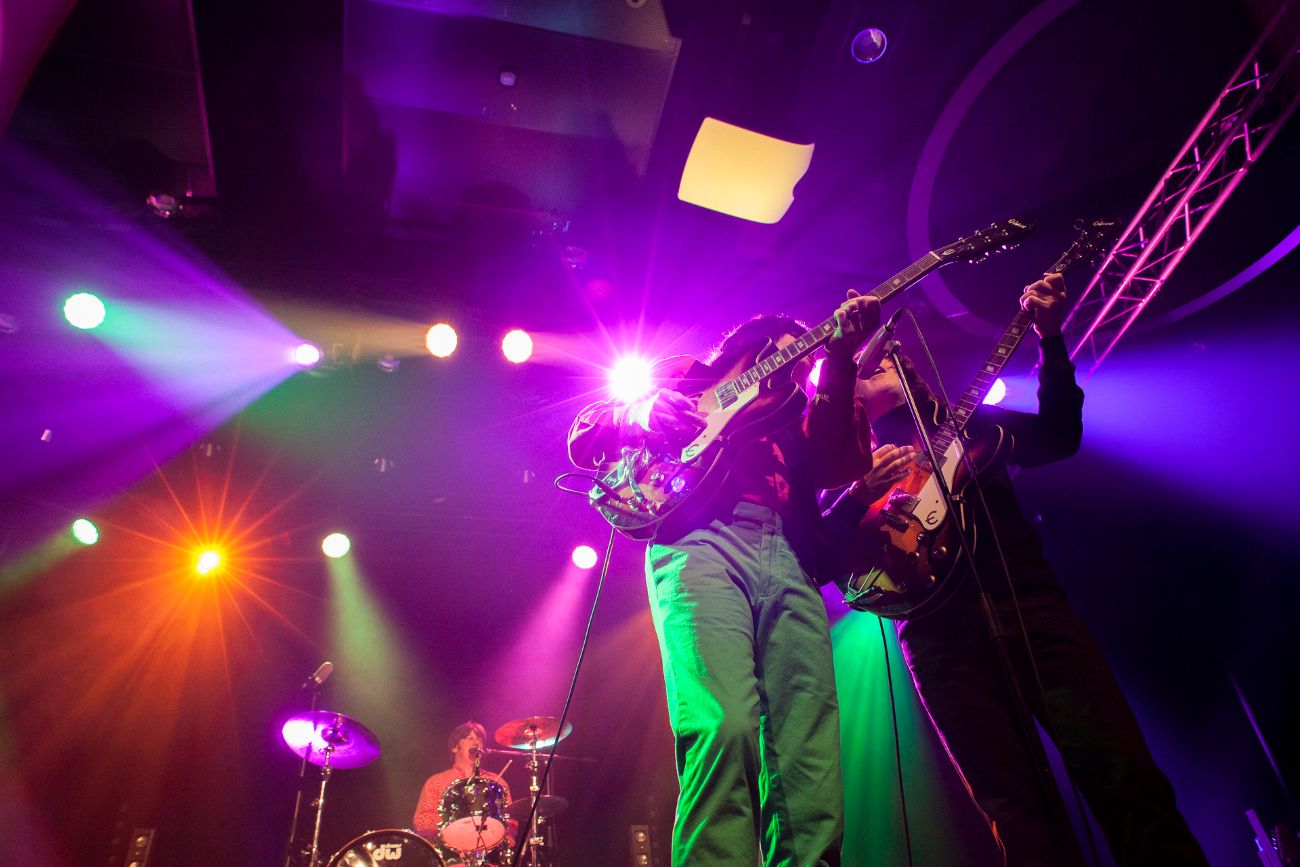 To gitarister spiller sammen på en opplyst scene med en trommeslager i bakgrunnen under fargerike konsertlys.