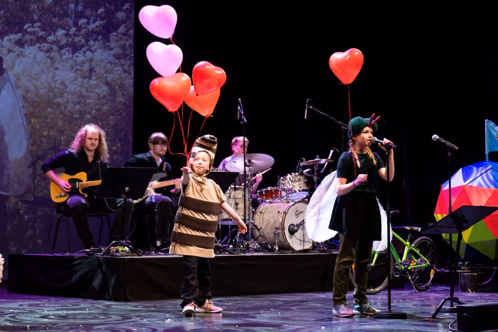 Barn kledd ut som bie og et annet barn kledd ut som flue holder hjerteformede ballonger på en scene med et band som spiller bak dem.