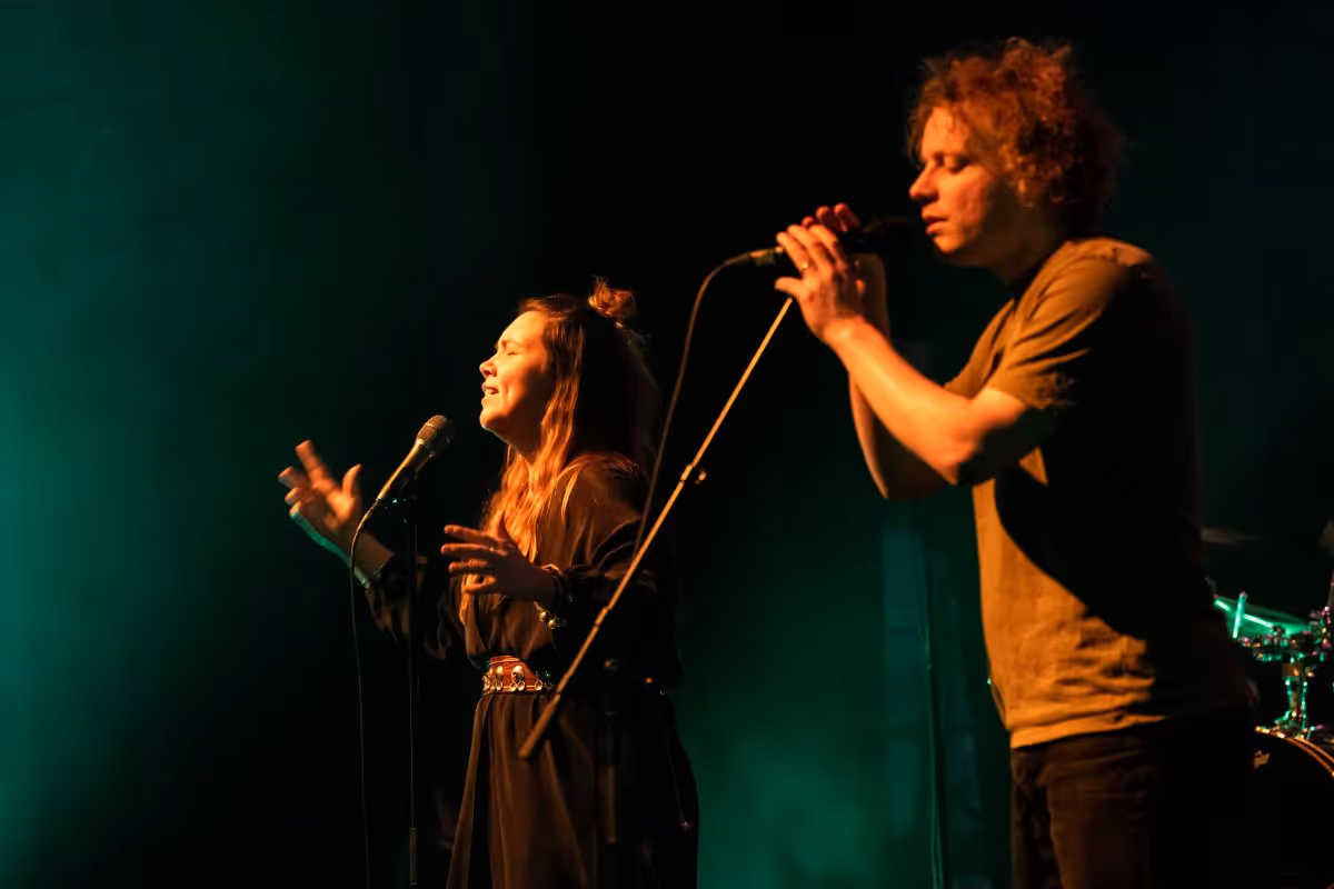 To musikere, en kvinne og en mann, synger med mikrofoner på en mørk scene med grønn bakgrunnsbelysning.
