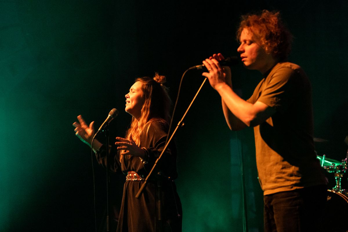 To musikere, en kvinne og en mann, synger med mikrofoner på en mørk scene med grønn bakgrunnsbelysning.