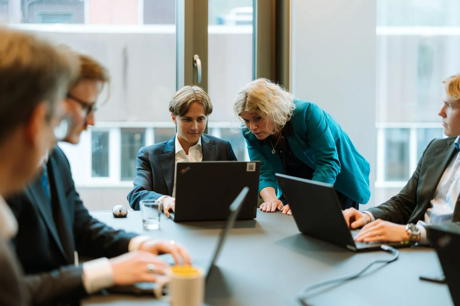 Fem unge voksne i formelle klær som samarbeider rundt et bord med bærbare datamaskiner i et kontormiljø.