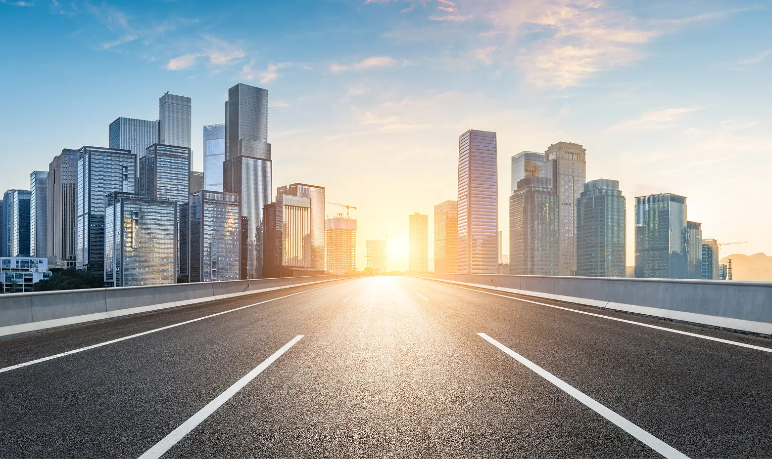 Empty highway leading toward a modern city skyline with high-rise buildings at sunrise.
