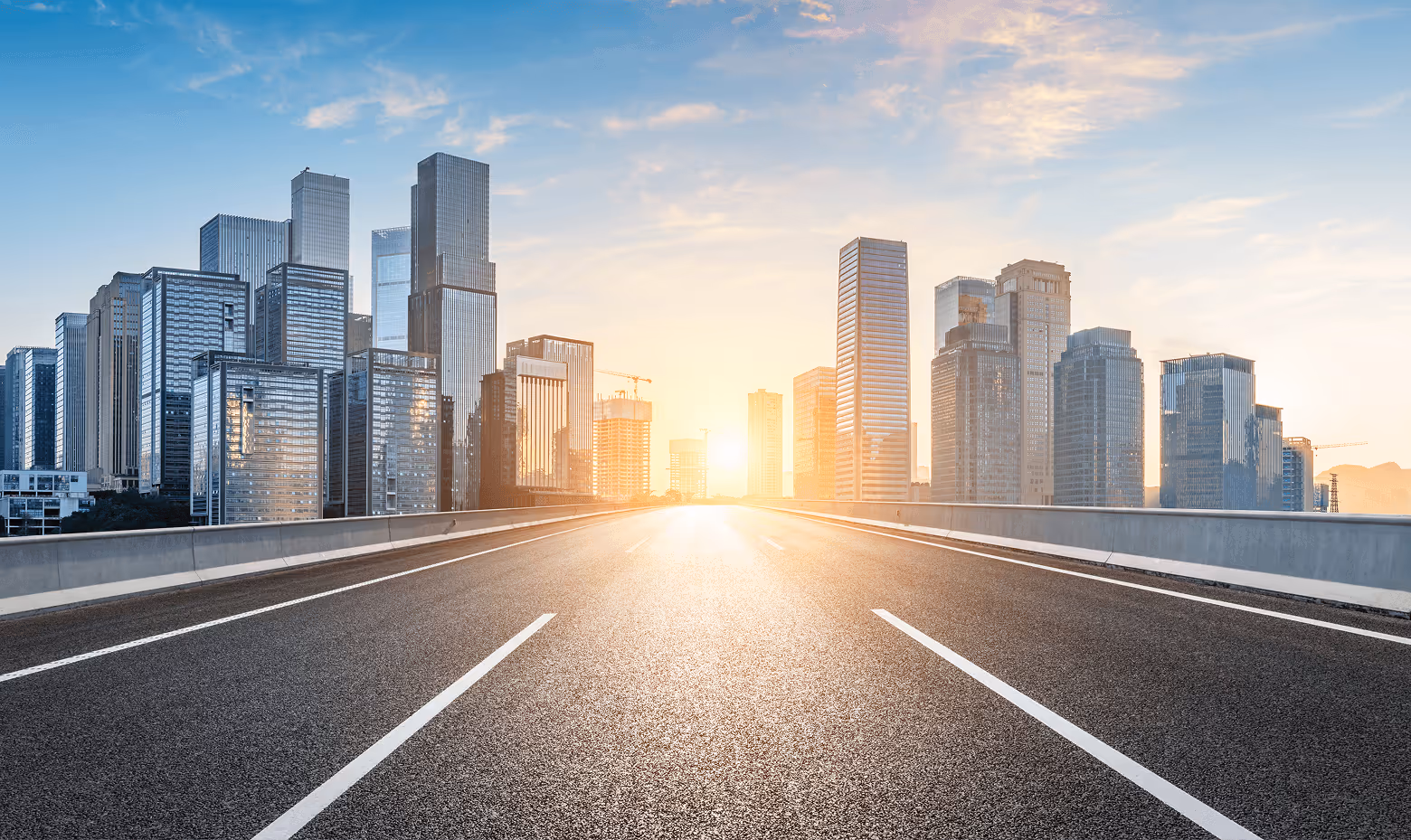 Empty highway leading towards a city skyline with modern skyscrapers at sunrise.