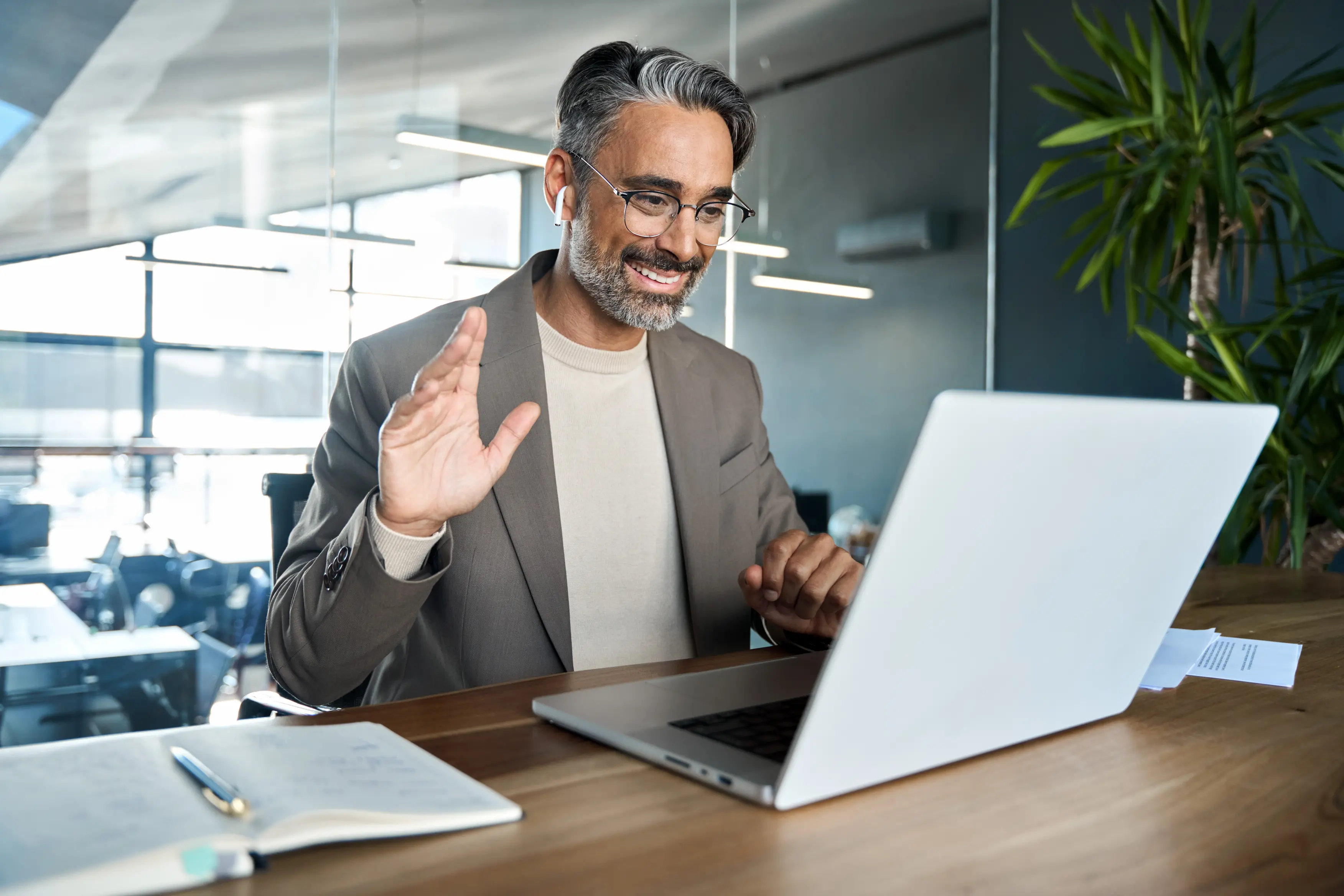 Smiling man with gray hair and glasses waving during a video call on a laptop at a wooden desk.