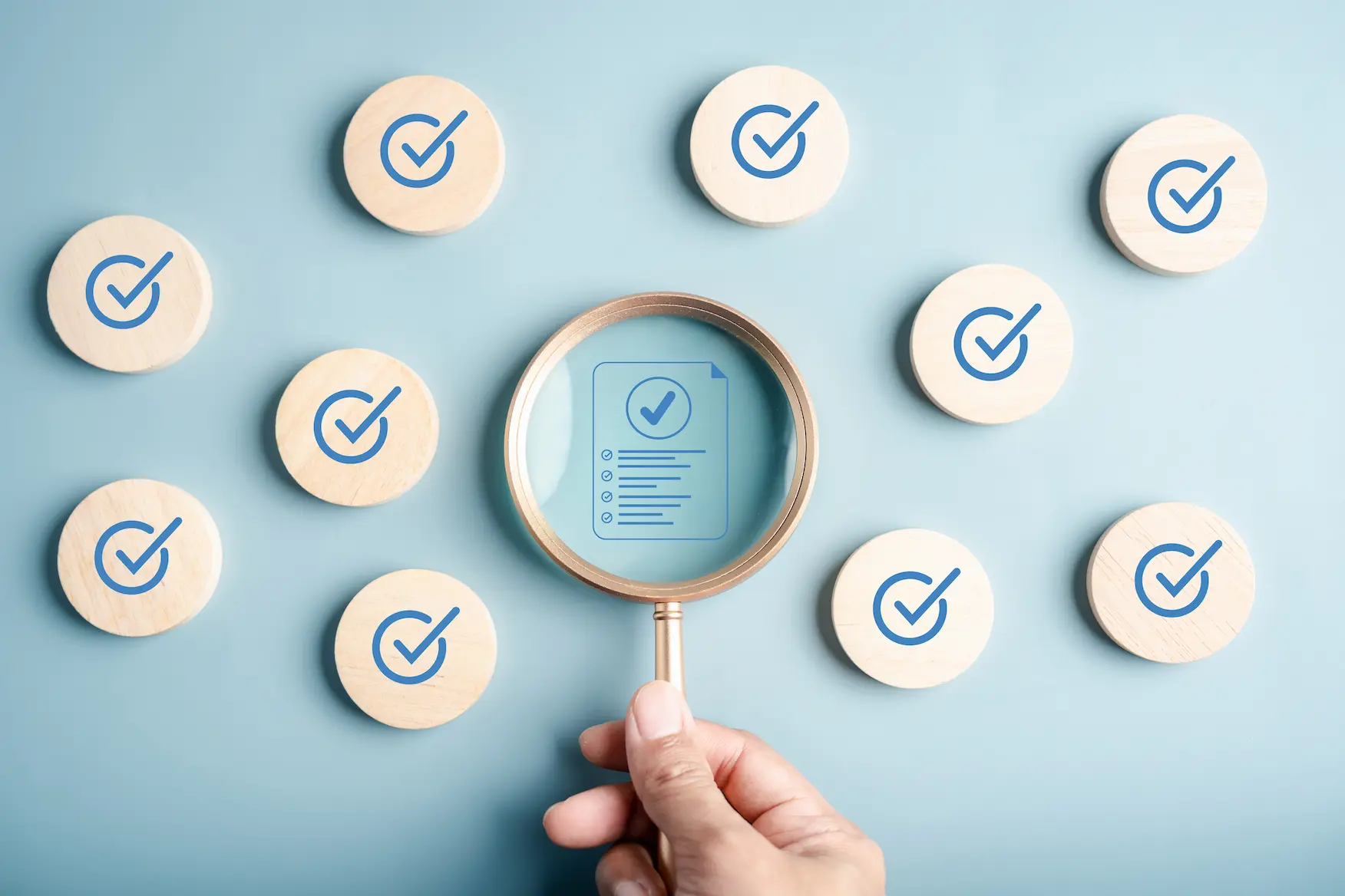 Hand holding a magnifying glass focusing on a checklist icon among wooden disks with blue checkmarks on a light blue background.