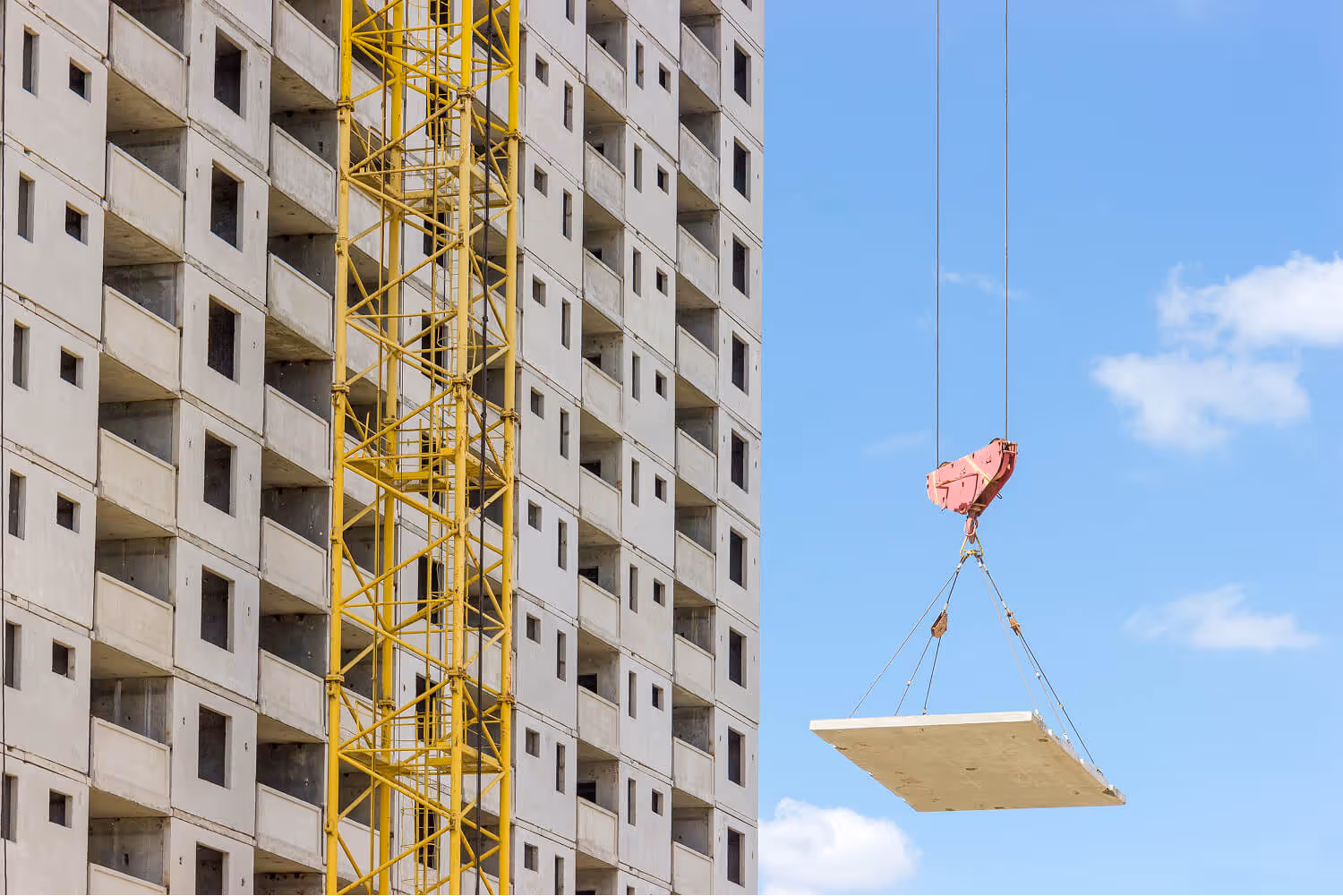 Yellow construction crane lifting a large concrete slab near an unfinished multi-story building under a blue sky.