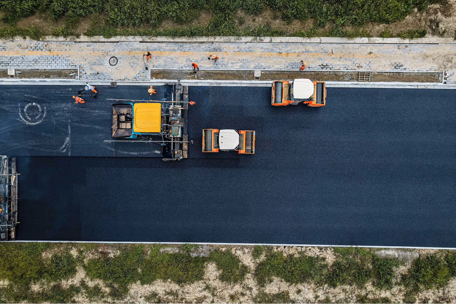 Aerial view of road construction with workers and machinery paving and rolling fresh asphalt.