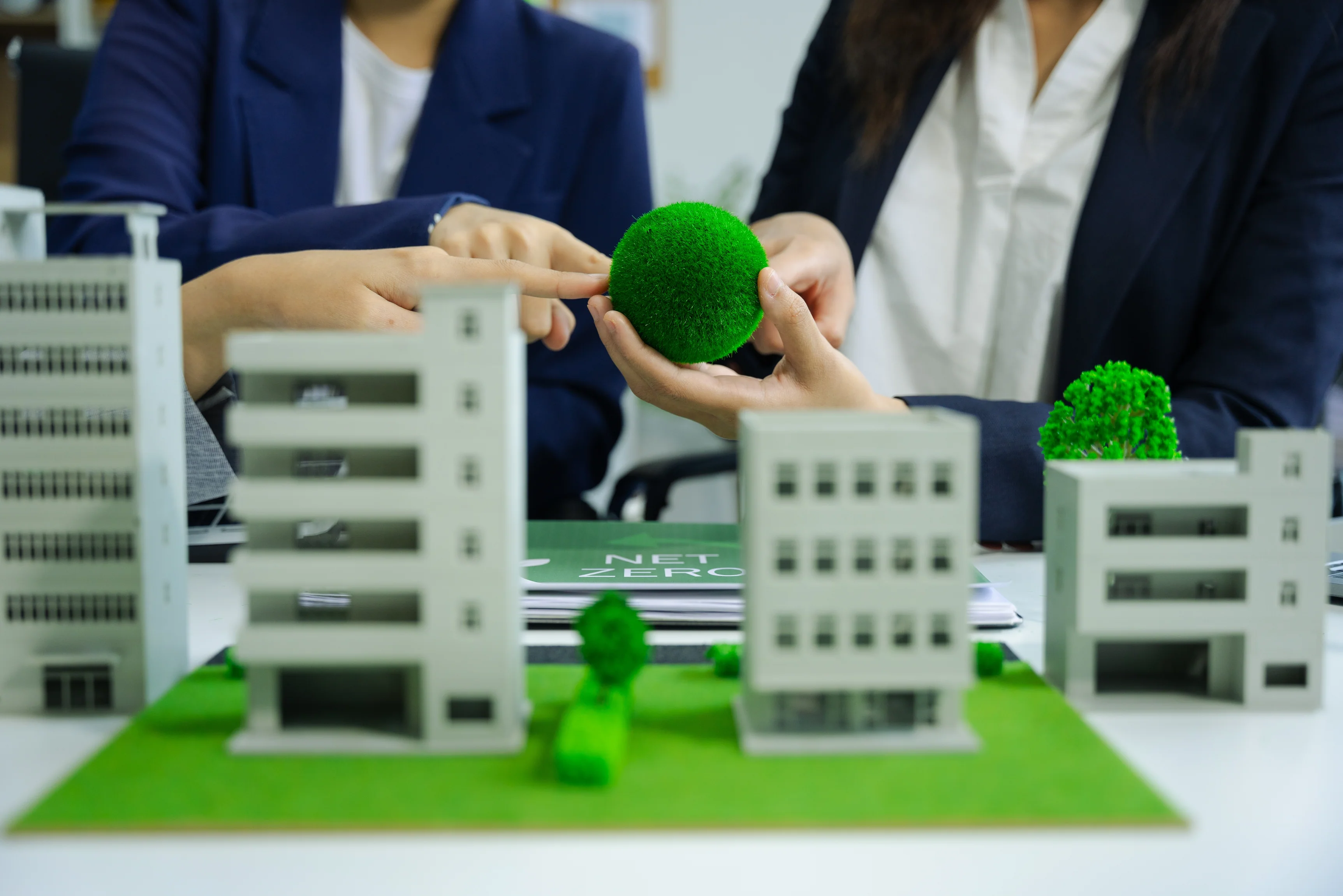 Two people in business attire hold a green moss ball above a miniature model of buildings and greenery, with a net zero booklet in the background.