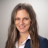 Lächelnde Frau mit langem braunen Haar, trägt einen dunklen Blazer und ein weißes Hemd vor hellem Hintergrund.