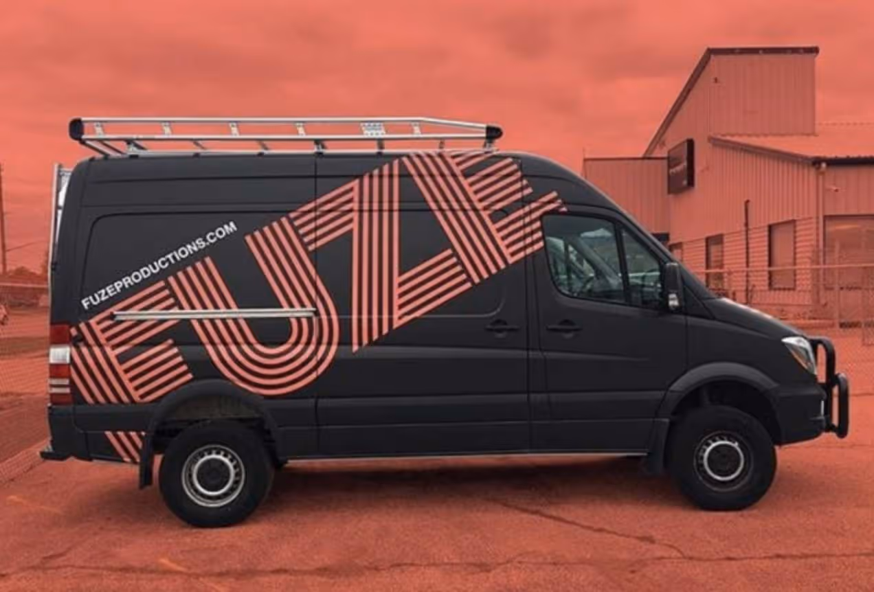 Black sprinter van with large red striped 'FUZE' text and website fuzeproductions.com on its side, parked in front of a red industrial building.