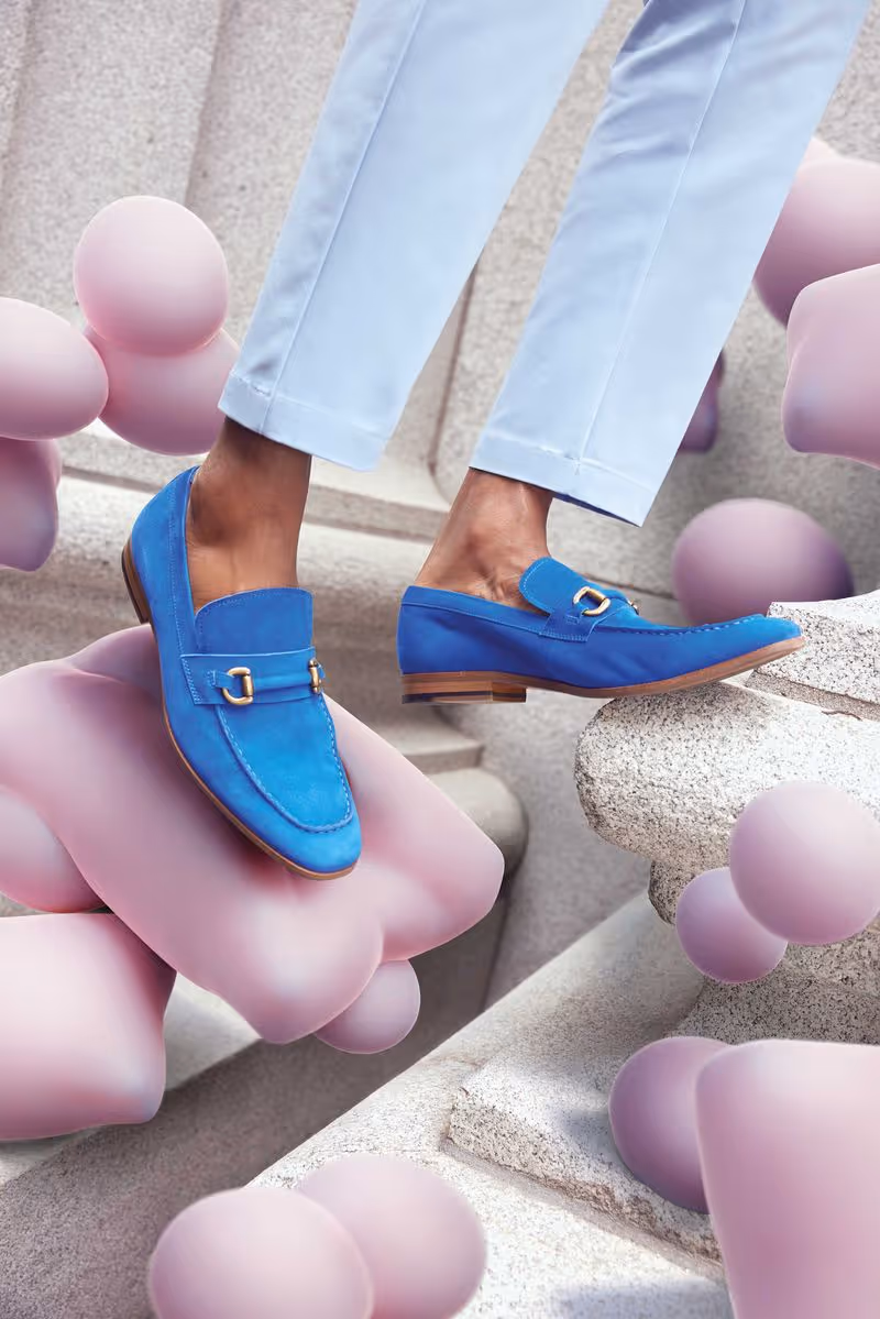 Person wearing bright blue loafers with gold buckles and light blue pants stepping on a stone step surrounded by abstract pink shapes.