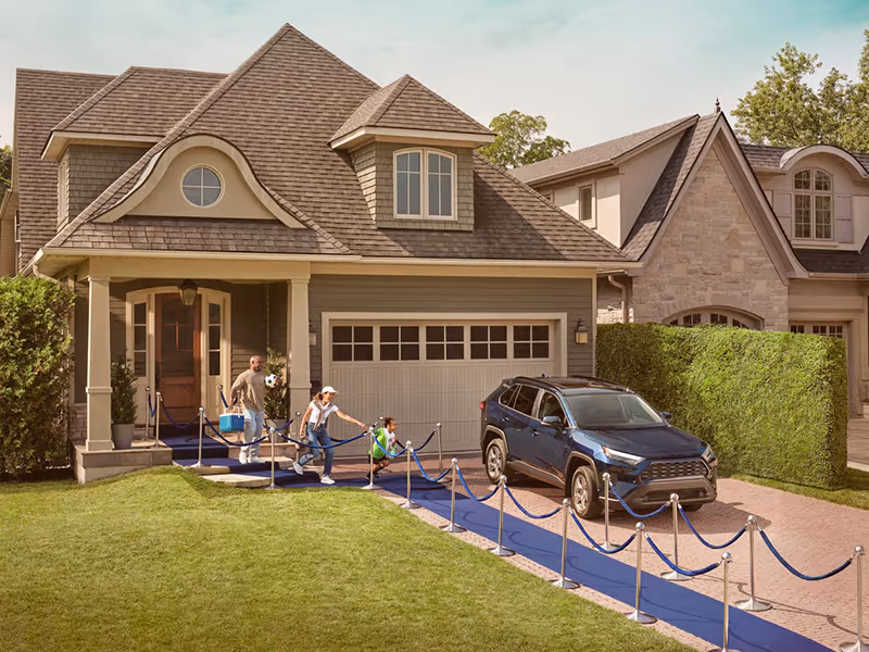 A family with children runs excitedly along a blue carpet leading from the driveway of a suburban house to the front door, with a parked dark blue SUV on the driveway.