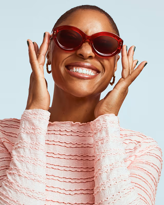 Smiling woman wearing large red sunglasses and a pink textured top against a light background.
