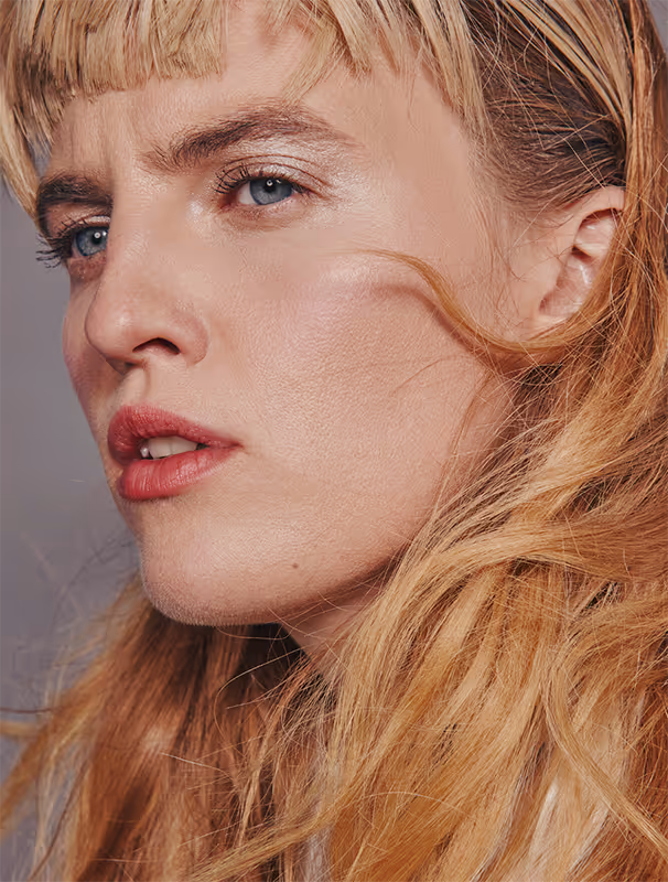 Close-up of a woman with blonde hair, blue eyes, pink lipstick, and a serious expression looking to the left.