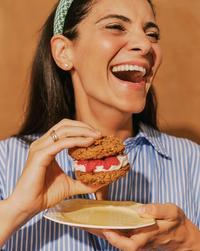 Woman with a green headband and striped shirt laughing while holding a raspberry and cream cookie sandwich above a yellow plate.