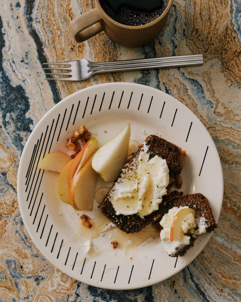 Plate with slices of pear, dark bread topped with ricotta cheese and honey, next to a fork and a coffee cup on a marbled tabletop.