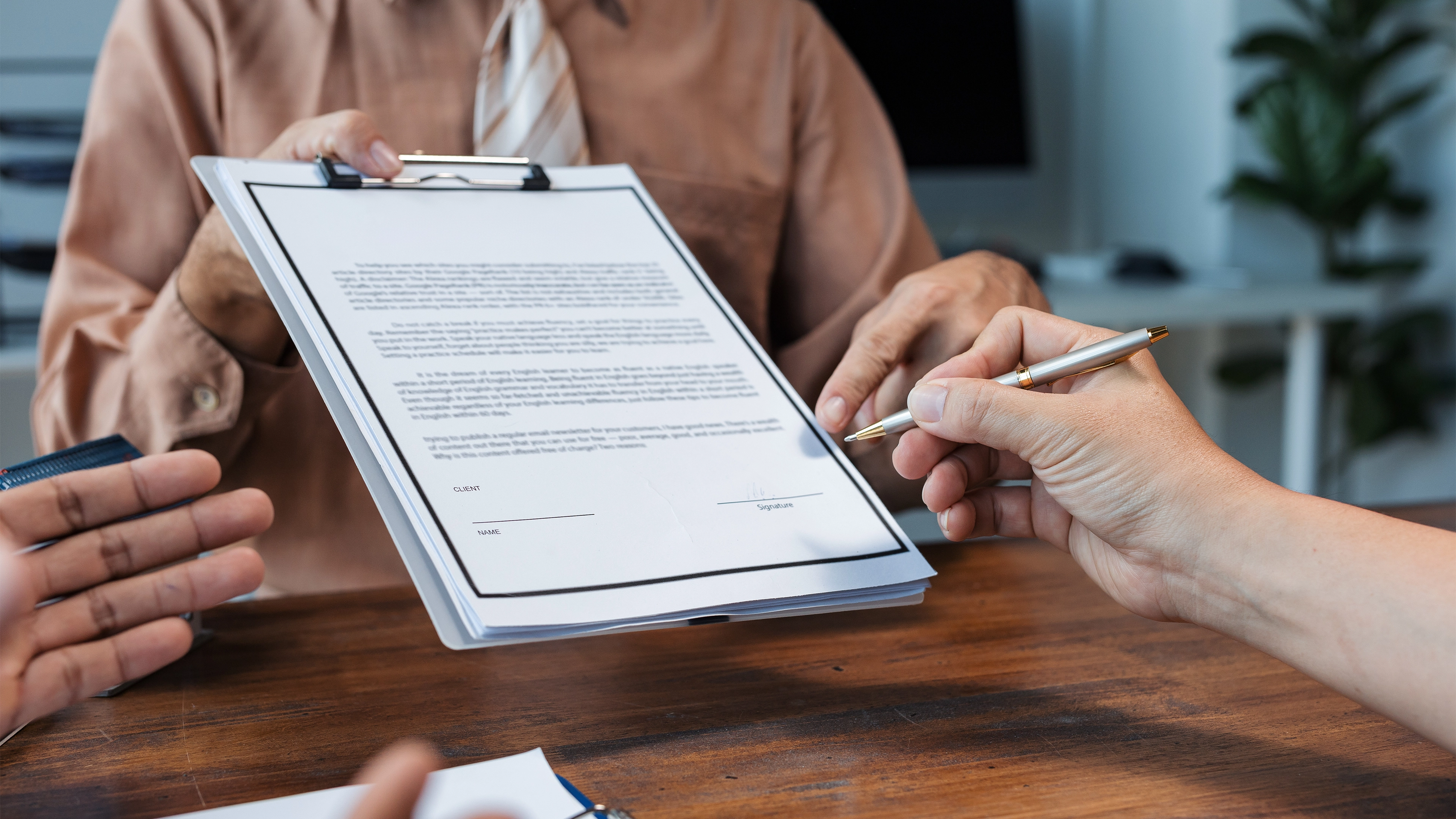 A person holding a clipboard with a document while another person prepares to sign it with a pen.