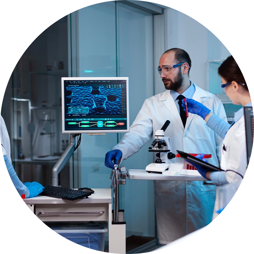 Two scientists in lab coats and safety glasses examining a blood sample with a microscope and computer displaying molecular data.