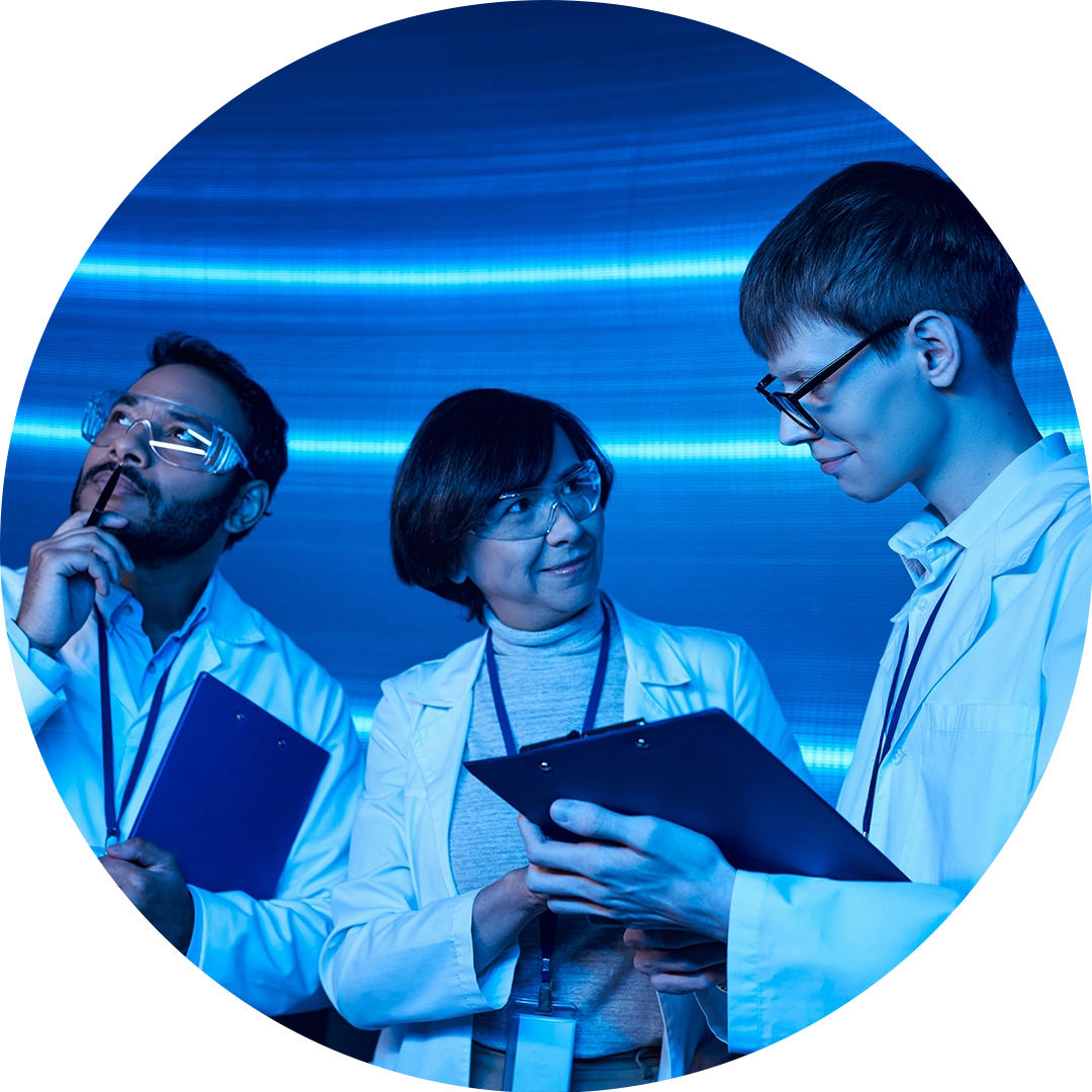 Three scientists in lab coats and safety glasses discussing notes on clipboards under blue lighting.