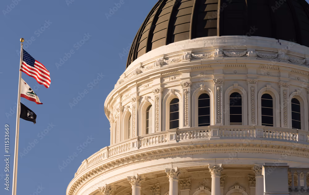 Close-up of the ornate white dome of a government building with American, California, and POW flags flying on a flagpole against a clear blue sky.