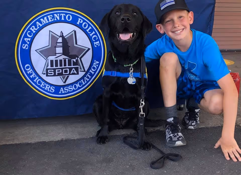 A young boy in a blue t-shirt poses beside a black Labrador retriever, both smiling. They sit in front of a backdrop featuring the Sacramento Police Officers Association logo.