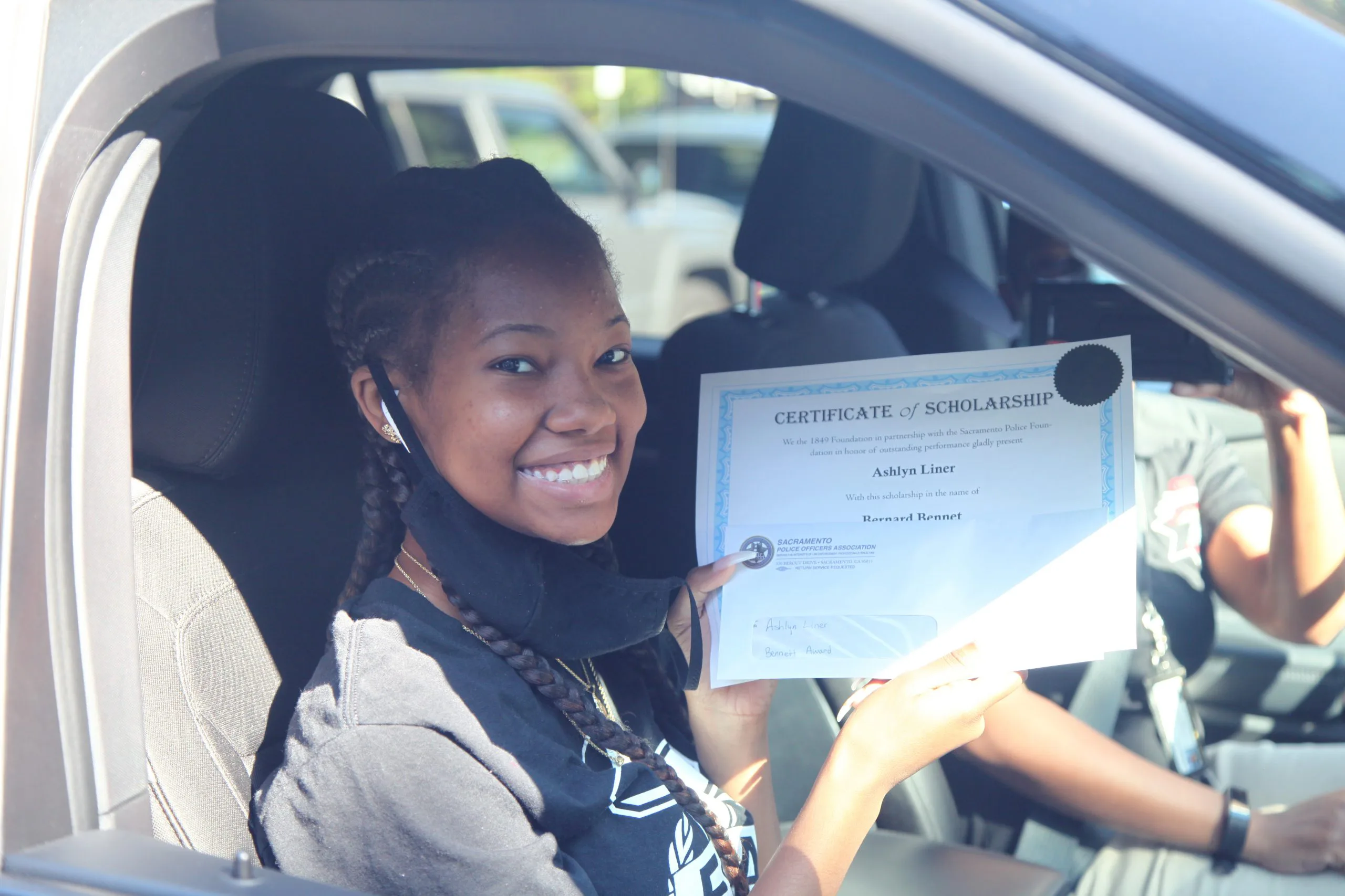 A young woman holding a scholarship certificate while sitting in a car. She is smiling and wearing a black shirt and a face mask. The interior of the vehicle and another person in the background are partially visible.
