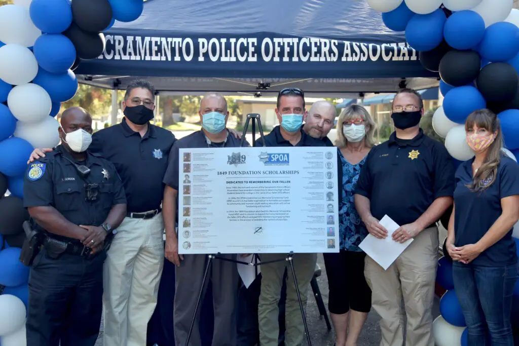 A group of seven police officers standing together in front of a banner for the Sacramento Police Officers Association. They are holding a document and wearing masks. Colorful balloons are in the background.