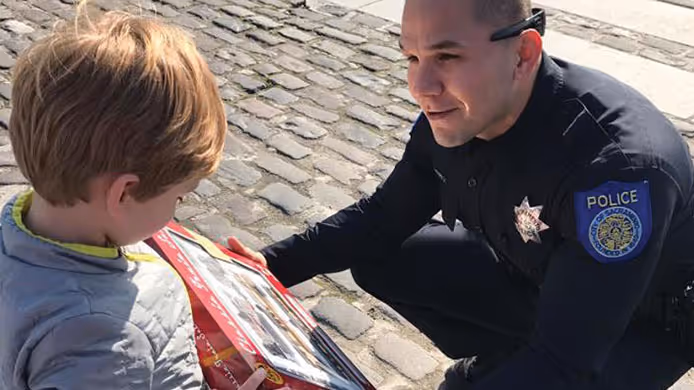 Police officer kneeling down and showing a book to a young boy on a cobblestone street.