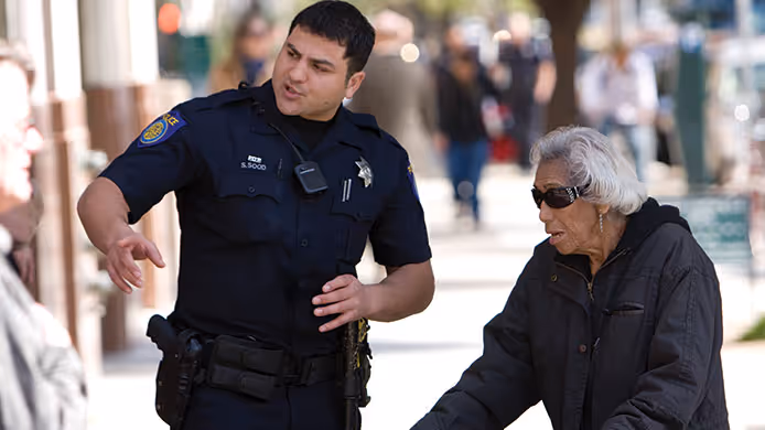 Police officer talking and gesturing to an elderly woman wearing sunglasses and a black jacket on a city sidewalk.