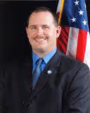 Man in dark suit and blue tie smiling in front of an American flag.