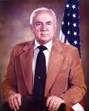 Portrait of an older man with white hair wearing a brown jacket and holding a dark hat, standing in front of an American flag.