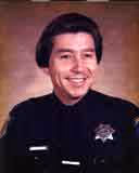Portrait of a young man in a dark uniform with a badge, smiling against a brown background.